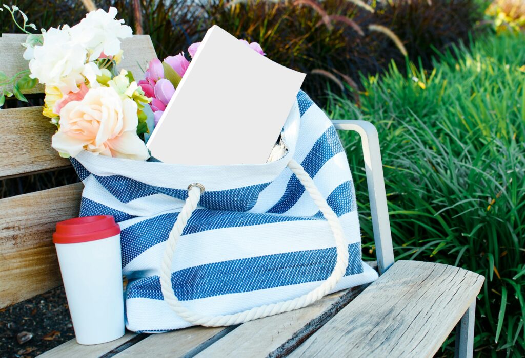 A striped beach bag filled with flowers and a book on a wooden bench outdoors.